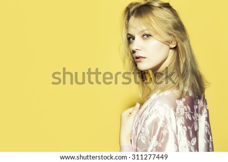 One beautiful young sensual pensive woman with blond hair in satin pink dressing gown in soft flower pattern pastel colour standing on yellow studio background copyspace, horizontal picture