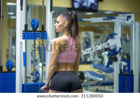 Back view of strong young attractive lady with straight beautiful body and bottom in pink and grey sportswear training in gym making weight-lifting exercises standing indoor, horizontal picture