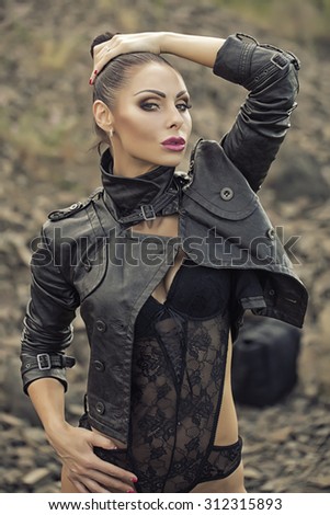 Pensive sexy brunette young wild woman with bright make-up and ponytail in black leather biker jacket and erotic lace lingerie looking forward outdoor on natural background, vertical picture