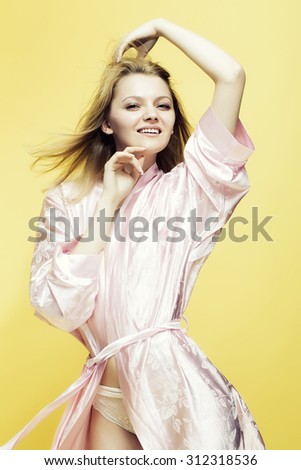 Blond beautiful young lovely smiling lady in satin pink dressing gown in soft flower pattern pastel colour pulling belt touching hair with hands standing on yellow studio background, vertical picture