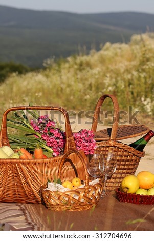 Picnic set of orange carrot green onion vegetable marrow pink flowers bouquet yellow ripe apples red wine bottle and empty glasses in baskets sunny day outdoor on natural background, vertical photo