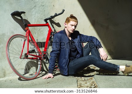 One cute red haired young fashionable unshaven stylish man in blue jacket sitting outdoor in street sunny day near bycicle leaning on grey wall, horizontal picture