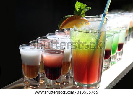 Many beautiful alcoholic shot cocktails in drinking glasses and one long beverage with straws mint and orange green brown red and white standing in row on black studio background, horizontal picture