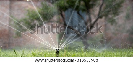 Closeup of one mechanical automatic irrigation system sprayed cold water as fountain on fresh green grass outdoor with tree and house on natural background, horizontal picture