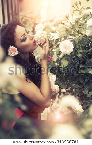 Pretty tender young woman with bright fashion makeup flower in brown curly hair shouting her eyes wearing dress with bare shoulders smelling holding rose posing near bush outdoor in park, vertical