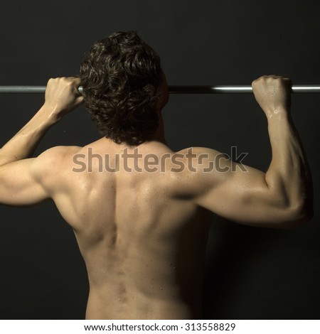 Back view of one young muscular sexy attractive athletic strong male with brunette curly hair doing chin-ups on metal bar with bare beautiful wet body on black studio background, square picture