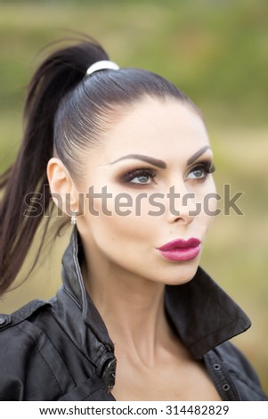 Portrait of cool pretty brunette young thoughtful woman with bright make-up and ponytail in black leather biker jacket looking away outdoor on green natural background, vertical picture