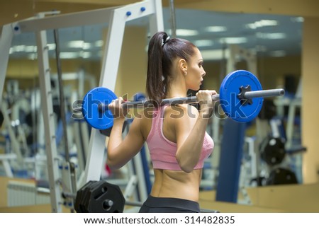 Back view of strong muscular young brunette sexual woman with straight beautiful body and bum in sportswear training in gym holding bar making weight-lifting exercises indoor, horizontal picture