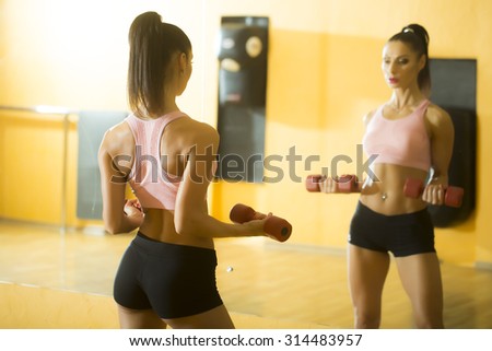 Back view of brunette sexual young slim woman with bright make-up and ponytail hairstyle training in gym in sportswear holding dumbell standing near mirror and reflecting, horizontal picture