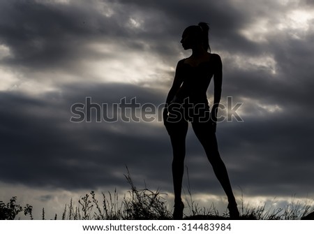Full length silhouette view of young woman with straight slim beautiful body standing outdoor in twilight on grey cumulus rainy clouds and moody sky natural background copyspace, horizontal picture