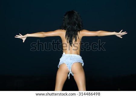 Back view of slim attractive sexual young girl with long brunette hair straight body and bottom in jeans shorts standing with raised hands in dusk on blue sky background outdoor, horizontal picture