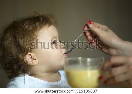 Nice little adorable toddler boy with hazel eyes and brown curly hair dressed in white t-shirt eating soup from glass bowl with spoon holding by mother on grey background indoor closeup, horizontal
