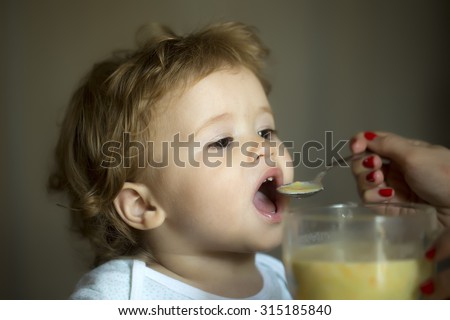 Little cute healthy hungry child boy with brown hair and hazel eyes dressed in t-shirt with open mouth eating soup from bowl with spoon holding by mother on grey background indoor closeup, horizontal