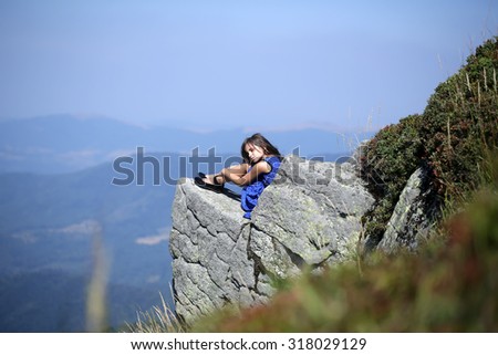 Pretty brunette pensive girl with long hair in blue lace dress sitting on stone rock cliff in beautiful landscape sunny day outdoor on natural blue sky background, horizontal photo