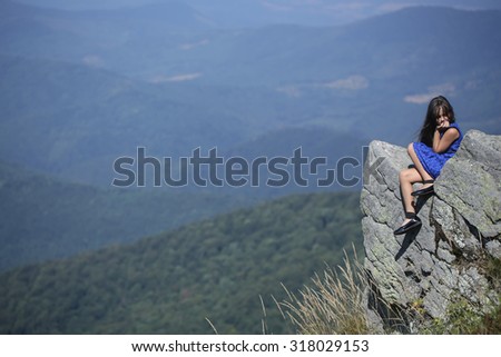 Pretty brunette small girl with long hair in blue lace dress sitting and thinking on stone rock cliff with beautiful landscape sunny day outdoor on natural blue sky background copyspace, horizontal