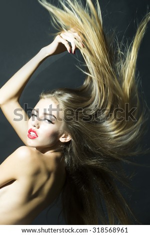 Portrait of one sensual young female model with long lush hair fluttering in wind naked back and red lips looking forward standing with raised hand in studio on black background, vertical picture