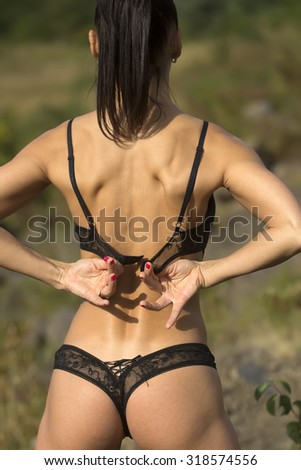 Back view of one young sexual brunette woman in black lace erotic lingerie with straight body and buttocks holding bra with hands standing outdoor sunny day on natural background, vertical picture