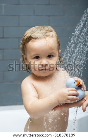 One beautiful happy baby boy with wet blonde curly hair in shower holding rubber toy of blue duckling standing under water indoor looking forward on grey background, vertical picture