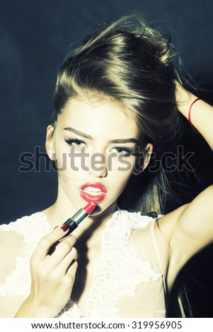 Portrait of fashionable pretty female model with lush hair putting on red lipstick on lips in white clothes from lace looking forward on black studio background closeup, vertical picture