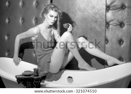 One beautiful sexy sensual young woman with bright makeup and curly hair in dress near column holding coffee cup sitting on bath tub in studio on leather black background, horizontal picture