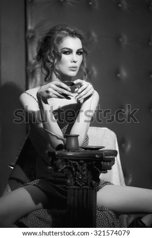 One beautiful sexy sensual young woman with bright makeup and curly hair in dress near column holding coffee cup sitting in studio on leather black background, vertical picture
