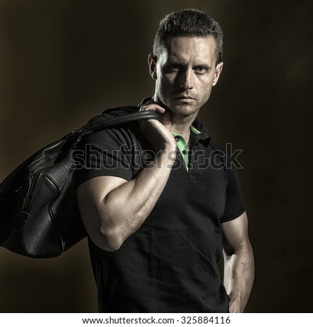 One handsome young muscular sexual business man in t-shirt standing in studio looking forward holding big leather hand bag on black background, square picture