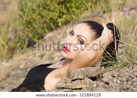 Sensuality young brunette lady with bright makeup and hair tail in black body lying on stony slope with hand behind head on natural background, horizontal picture