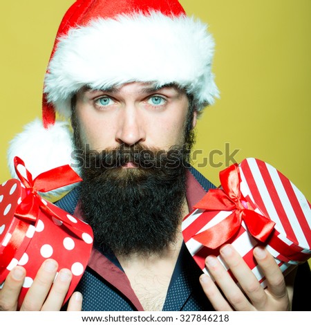 Portrait of one senior new year man with long black beard in shirt and red santa claus hat holding two wrapped present boxes in hands for christmas standing in studio on yellow background, square