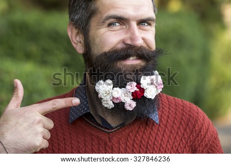 Closeup portrait of one handsome smiling man with long black beard showing on small many white and pink flowers in red sweater looking forward sunny day outdoor on natural green background, horizontal