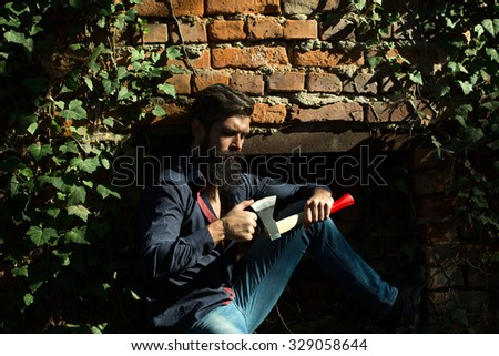 One handsome strong stylish male logger of young man with long lush black beard and moustache in shirt holding wooden axe standing near brick wall outdoor, horizontal picture