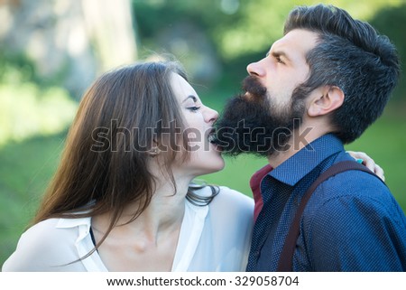 Portrait closeup view of beautiful funny young pair of woman biting man with long lush black beard standing sunny day outdoor on green grass on natural backdrop, horizontal photo