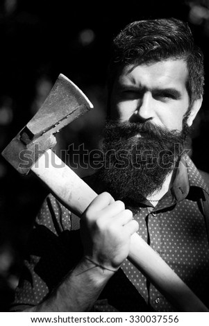 Portrait of one handsome strong stylish male logger of young serious man with long lush beard and moustache in shirt holding wooden axe standing outdoor on natural background black and white, vertical