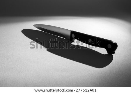 Sharp metal chef knife with a black handle on a white surface with a back light casting a shadow of the knife. It has a spooky feel.
