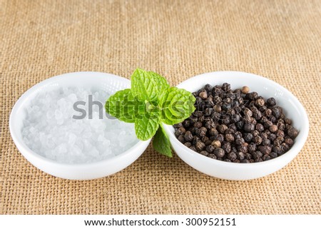 Two small white porcelain bowls, one filled with black pepper corns and one with white sea salt on a brown hessian mat with green mint leaves.