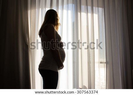 Silhouette of a pregnant woman holding her belly looking out a window with curtains and morning sunlight streaming in.