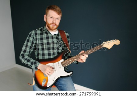 Young red hair man with red beard in plaid shirt playing guitar on dark background