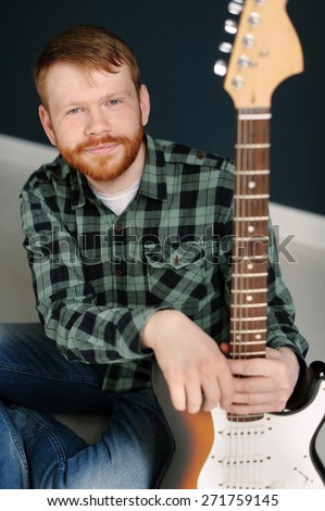 Young red hair man with red beard in plaid shirt holding a guitar on dark background