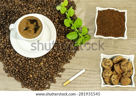 Cup of fresh coffee, coffee beans,  fresh ground coffee, cookies with chocolate, cigarette and mint leaves on wooden table