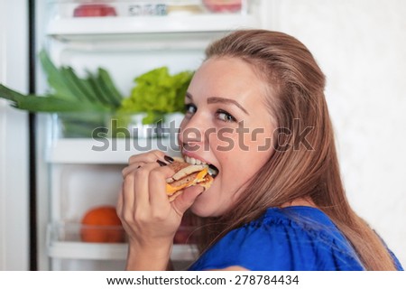 young losing weight woman bites off a Burger on the background of the fridge and food