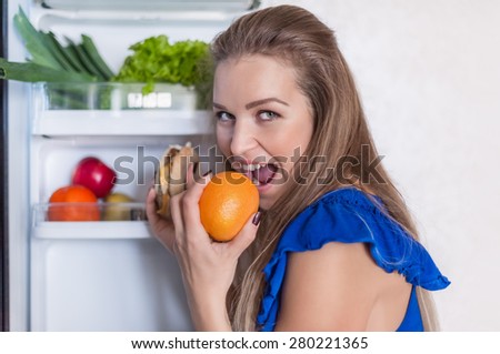young woman near open fridge-orange and cheeseburger in hand