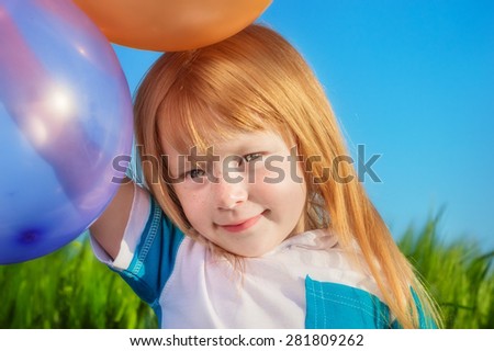 cute redheaded girl with freckles smiling and holding balloons on a background of green grass and blue sky