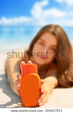 Young woman smiling while relaxing on beach and holding sunscreen bottle in her hands