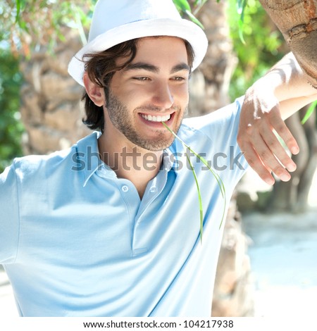 Young man wearing hat and casual clothes in sunny day. Leaning on palm tree at summer park