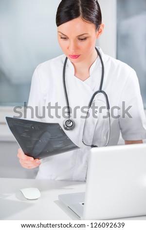Portrait of a female doctor holding ultrasound results and using her laptop computer at clinic