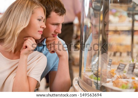 Couple shopping at the supermarket, standing near glass showcase and thinking what to buy