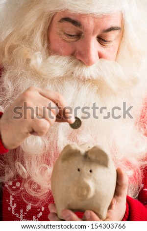 Authentic Santa Claus holding piggy bank and putting golden coin inside, Christmas budget concept