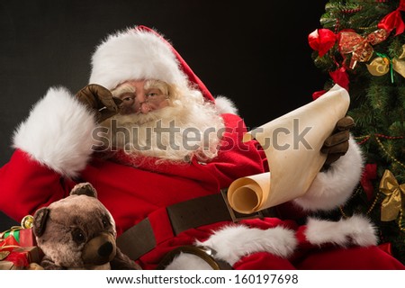 Portrait of happy Santa Claus sitting at his room at home near Christmas tree and big sack and reading Christmas letter or wish list