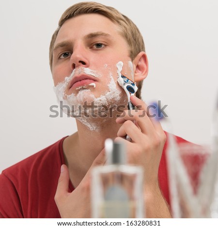 Young man shaving in the bath. He is passing the razor for the beard while  looks at the mirror