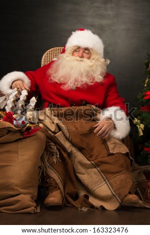 Santa Claus with a huge bag full of Christmas presents sitting in a comfortable chair near the Christmas tree at home