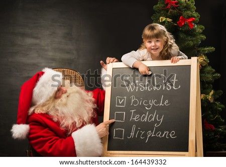 Santa Claus with child sitting near chalkboard with wish list and checking it